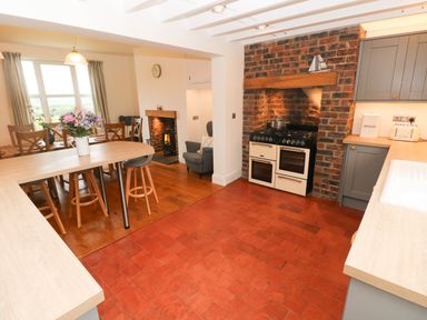 A kitchen and dining area with a stove in a brick alcove a dining table with chairs and flowers a small fireplace and an armchair at Copybush Cottage in Lawrenny