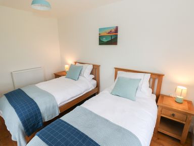 A bedroom with two single beds with blue blankets and cushions two wooden bedside tables with lamps and a landscape painting on the wall at Copybush Cottage in Lawrenny