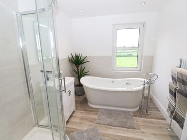 A bathroom with a freestanding bathtub a shower cubicle a sink a potted plant towels on a rack and a window showing green fields at Copybush Cottage in Lawrenny