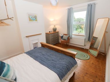 A bedroom with a wooden bed a chest of drawers a wicker chair a full-length mirror and a window with curtains at Copybush Cottage in Lawrenny