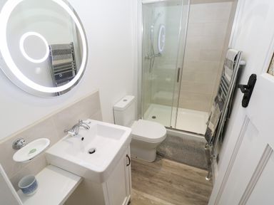 A bathroom with a sink cabinet, toilet, glass shower enclosure, towel rack, and round illuminated mirror at Copybush Cottage in Lawrenny