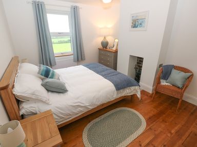 A bedroom with a wooden bed white bedding blue pillows wooden nightstand a small rug chest of drawers lamp and window with blue curtains at Copybush Cottage Lawrenny