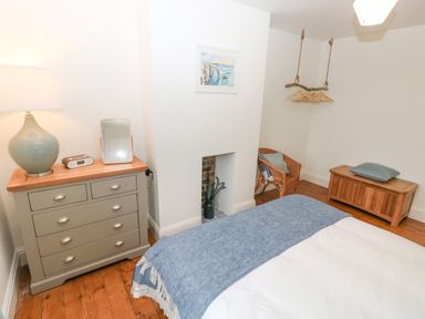 A bedroom with a bed covered by a blue blanket a gray chest of drawers with a lamp and clock a wicker chair a hanging hanger rack and a wooden storage bench at Copybush Cottage in Lawrenny