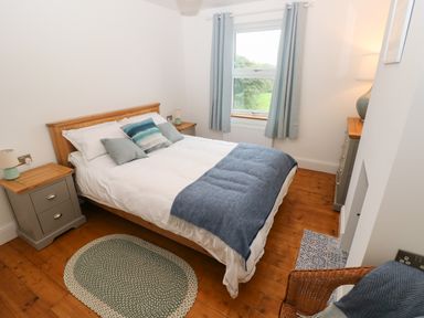 A bedroom with a double bed blue throw pillows bedside tables window with curtains and wooden floor at Copybush Cottage in Lawrenny