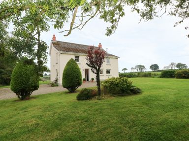 A white two-story house with a gravel driveway surrounded by a lawn with trees and shrubs at Copybush Cottage in Lawrenny