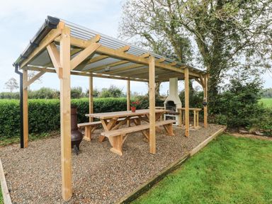 A wooden pergola with picnic tables and a white outdoor fireplace in a garden at Copybush Cottage in Lawrenny