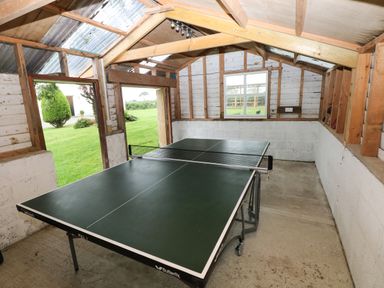 A room with a ping pong table inside a wooden structure with open doors and windows showing a grassy yard outside at Copybush Cottage in Lawrenny
