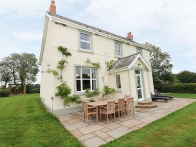 The exterior of a house with outdoor dining table and chairs on a patio and a lawn with trees at Copybush Cottage in Lawrenny