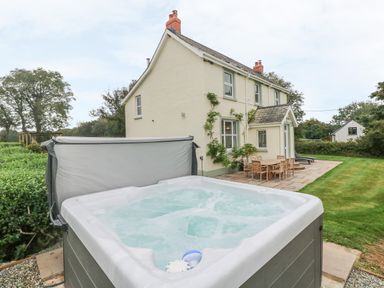 An outdoor hot tub with water bubbling beside a patio with a wooden table and chairs next to a two-story cream house surrounded by grass at Copybush Cottage in Lawrenny