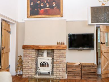 A room with a brick fireplace and a white stove inside it a wall-mounted television wooden crates labeled wereham school logs and a framed painting above the fireplace at The Old School in Downham Market