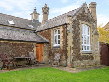 An outdoor patio area with wooden chairs and a table by a stone house with white windows and a wooden door at The Old School in Downham Market