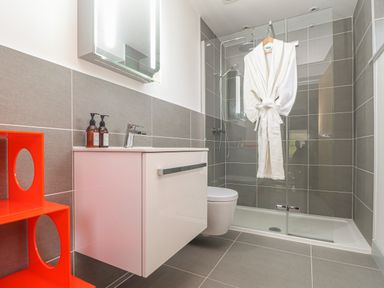 A bathroom with a white vanity cabinet with two soap dispensers a glass shower enclosure with a white robe hanging inside and an orange shelving unit at Idlewild 2 Wheal Turnavore in St Agnes