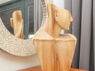 A wooden abstract bust sculpture on a table in front of a patterned round mirror and gray curtains at Idlewild 2 Wheal Turnavore in St Agnes