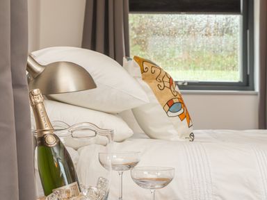 A bedroom with a white bed linens a decorative pillow a window a table lamp a bottle of champagne in an ice bucket and two champagne glasses at Idlewild 2 Wheal Turnavore in St Agnes