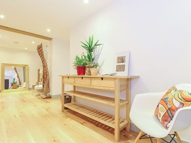 A hallway with a wooden console table with plants and a picture frame next to a white chair with a colorful cushion at Idlewild 2 Wheal Turnavore in St Agnes