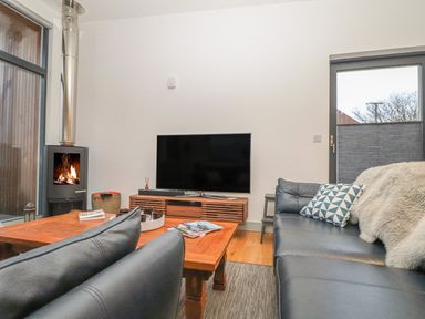 A living room with a black leather sofa a wooden coffee table a flat screen tv on a wooden stand and a wood burning stove at Idlewild 2 Wheal Turnavore in St Agnes