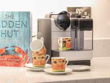 A coffee machine with four colorful cups and saucers on a kitchen counter next to a cookbook at Idlewild 2 Wheal Turnavore in St Agnes