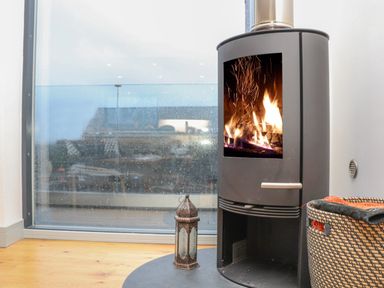 A wood burning stove with fire inside near a window with rain outside and a wicker basket with blankets at Idlewild 2 Wheal Turnavore in St Agnes