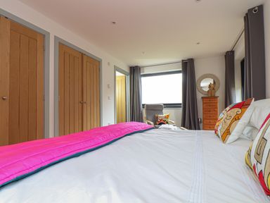 A bedroom with a bed with colorful pillows a pink blanket wooden doors a gray armchair with a pillow and a wooden chest with a round mirror at Idlewild 2 Wheal Turnavore in St Agnes