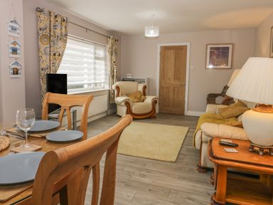 A living room with a dining table set with plates and glasses a cream armchair a cream sofa with pillows and a yellow blanket a wooden door and floral curtains at 2 The Close in Robin Hood'S Bay
