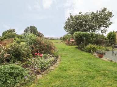 A garden with flower beds and a grassy path leading to outdoor wooden chairs and a table at 2 The Close in Robin Hood'S Bay