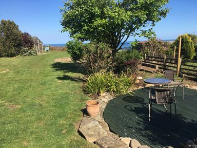A garden with a round wooden deck featuring a small table and two chairs with bushes trees and a greenhouse in the background at 2 The Close in Robin Hood'S Bay