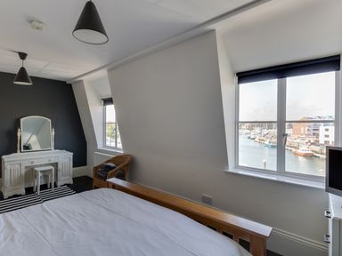 A bedroom with a wooden bed frame a vanity table a wicker chair and two windows overlooking a marina at Sailor's Watch in Weymouth