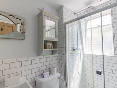 A bathroom with a shower area, white tiled walls, a wall cabinet with a mirror, a toilet with a roll of toilet paper, and a sink with a toothbrush holder at Sailor's Watch in Weymouth