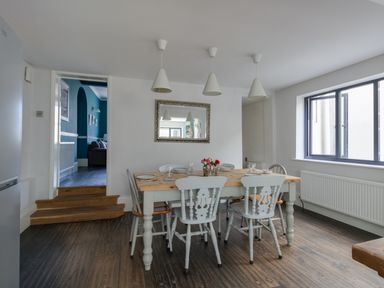 A dining room with a wooden table, six chairs, three pendant lights, and a window at Sailor's Watch in Weymouth