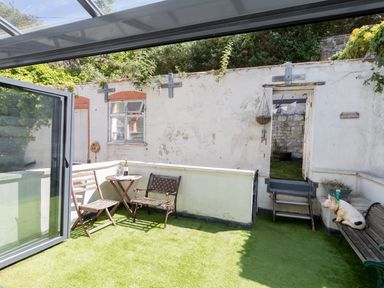 An outdoor patio area with artificial grass two chairs a small table a bench with a pig decoration and a doorway leading to a garden at Sailor's Watch in Weymouth