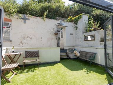 An outdoor patio with artificial grass two wooden benches a small round wooden table with a bottle and glasses and white walls at Sailor's Watch in Weymouth