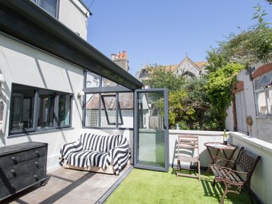 A patio area with a striped sofa a black chest a wooden chair a bench and a small table with wine glasses at Sailor's Watch in Weymouth