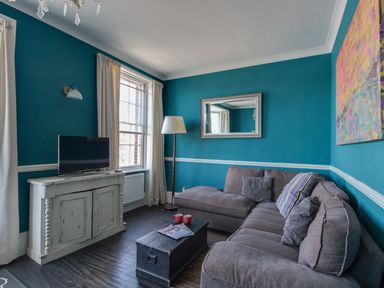 A living room with a gray sectional sofa a black trunk table a white cabinet with a TV and blue walls at Sailor's Watch in Weymouth