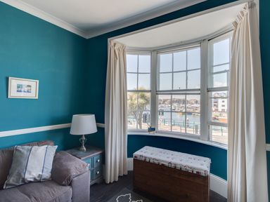 A living room corner with teal walls a gray sofa a side table with a lamp a window with white curtains and a cushioned wooden bench at Sailor's Watch in Weymouth