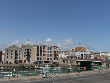 A street with a small white scooter and pedestrians next to a canal with boats and buildings along the water in Weymouth