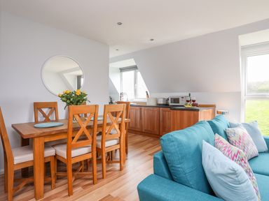 A dining area with wooden chairs and a table with flowers next to a kitchen with wooden cabinets and a blue sofa at Isallt Cove in Trearddur Bay