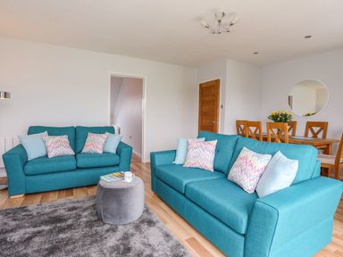 A living room with two blue sofas and a round gray ottoman next to a dining table with wooden chairs at Isallt Cove in Trearddur Bay