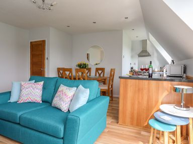 A living area with a turquoise sofa and multicolored pillows a wooden dining table with chairs and a round mirror on the wall a kitchen area with a wooden counter a microwave and wine glasses at Isallt Cove in Trearddur Bay