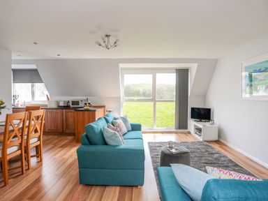 A living room with blue sofas a gray rug a small gray ottoman and a television on a white stand at Isallt Cove in Trearddur Bay