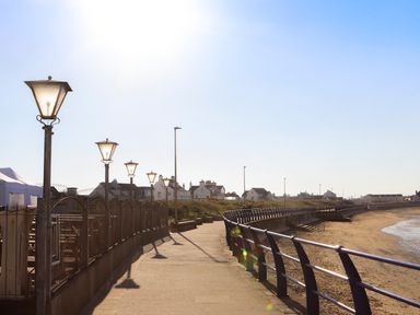 A curved promenade with street lamps and railings along a sandy beach with houses in the background at Isallt Cove in Trearddur Bay
