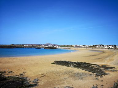 A sandy beach with rocks and calm water with houses in the background at Isallt Cove in Trearddur Bay