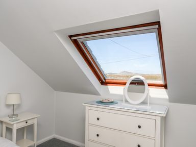 A white dresser with a round mirror and a plate under a skylight window next to a white nightstand with a lamp at Isallt Cove in Trearddur Bay
