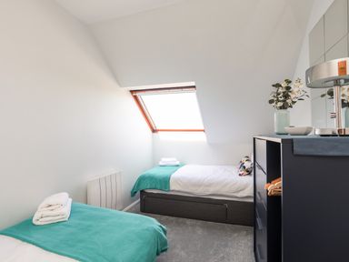 A bedroom with two single beds covered with teal blankets a chest of drawers with flowers and a lamp by a skylight window at Isallt Cove in Trearddur Bay