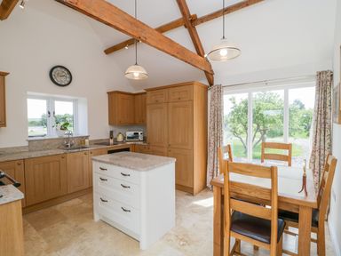 A kitchen with wooden cabinets a center island and a dining table with chairs next to a large window at Golden Valley Barn in Oldbury-On-Severn