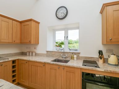 A kitchen with wooden cabinets a sink below a window a clock above the window a cooktop an oven a kettle and a knife block at Golden Valley Barn in Oldbury-On-Severn