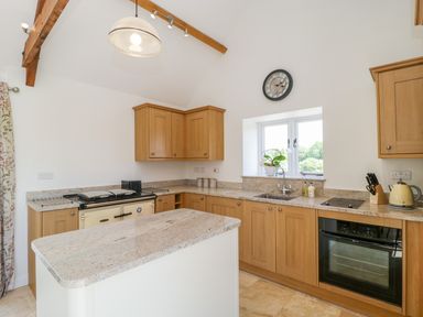A kitchen with wooden cabinets, a granite countertop island, an oven, stove, sink, and a window with a clock above at Golden Valley Barn in Oldbury-On-Severn