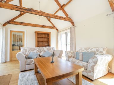 A living room with two floral sofas a wooden coffee table a wooden bookshelf exposed wooden ceiling beams and glass doors at Golden Valley Barn in Oldbury-On-Severn