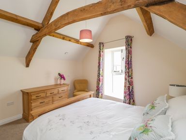 A bedroom with wooden beams a window with floral curtains a wooden dresser a yellow chair and a bed with floral pillows at Golden Valley Barn in Oldbury-On-Severn