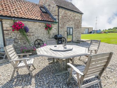 An outdoor patio with wooden chairs and a table next to a stone building with hanging flower baskets at Golden Valley Barn in Oldbury-On-Severn