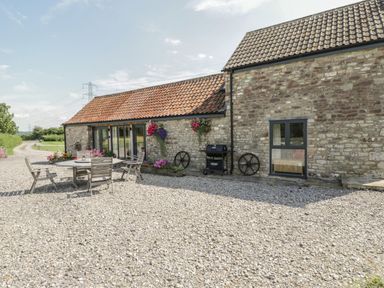 An outdoor patio area with a round table and chairs next to a stone wall with flowers and a barbecue grill at Golden Valley Barn in Oldbury-On-Severn